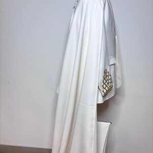 Puede incluir: Un blazer blanco con adornos geom&eacute;tricos dorados en el bolsillo y el hombro. El blazer tiene una larga capa fluida. La prenda se exhibe en un maniqu&iacute; sobre un fondo blanco.