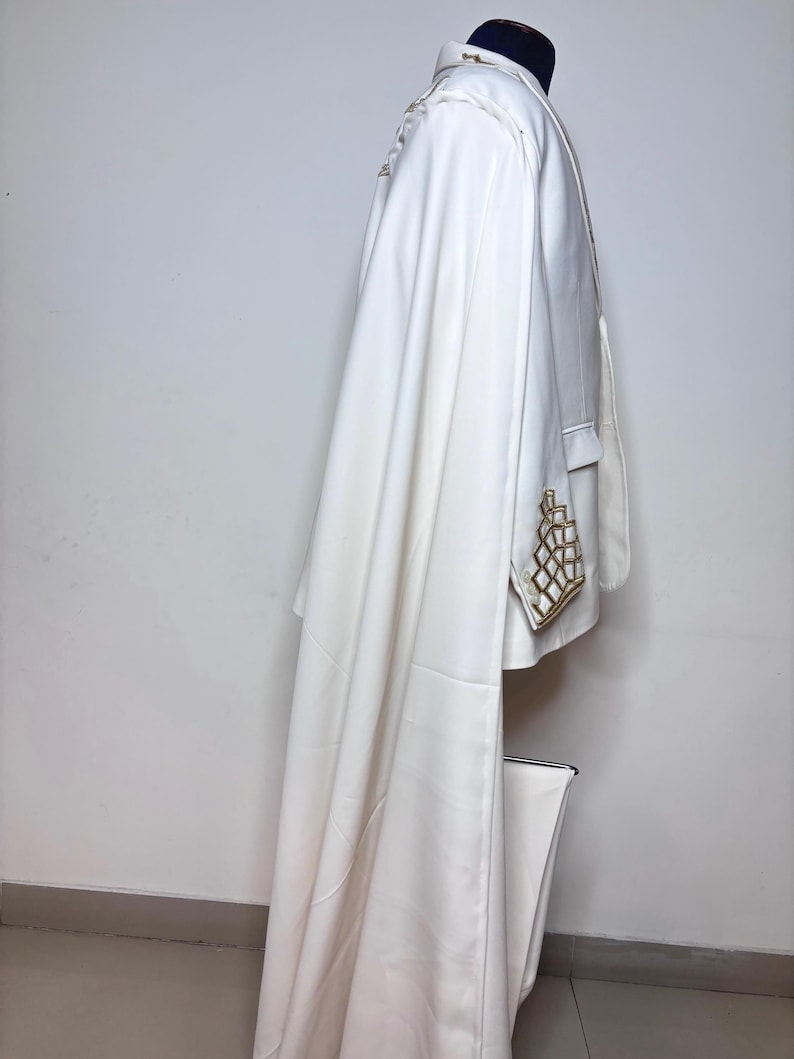 Puede incluir: Un blazer blanco con adornos geom&eacute;tricos dorados en el bolsillo y el hombro. El blazer tiene una larga capa fluida. La prenda se exhibe en un maniqu&iacute; sobre un fondo blanco.