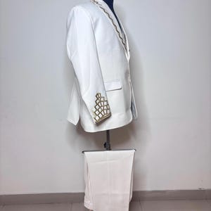 Puede incluir: Un traje blanco exhibido en un maniqu&iacute;. El traje incluye una chaqueta con bordados geom&eacute;tricos dorados en la solapa y los pu&ntilde;os, y pantalones a juego. El traje es entallado y parece estar hecho de una tela lisa.