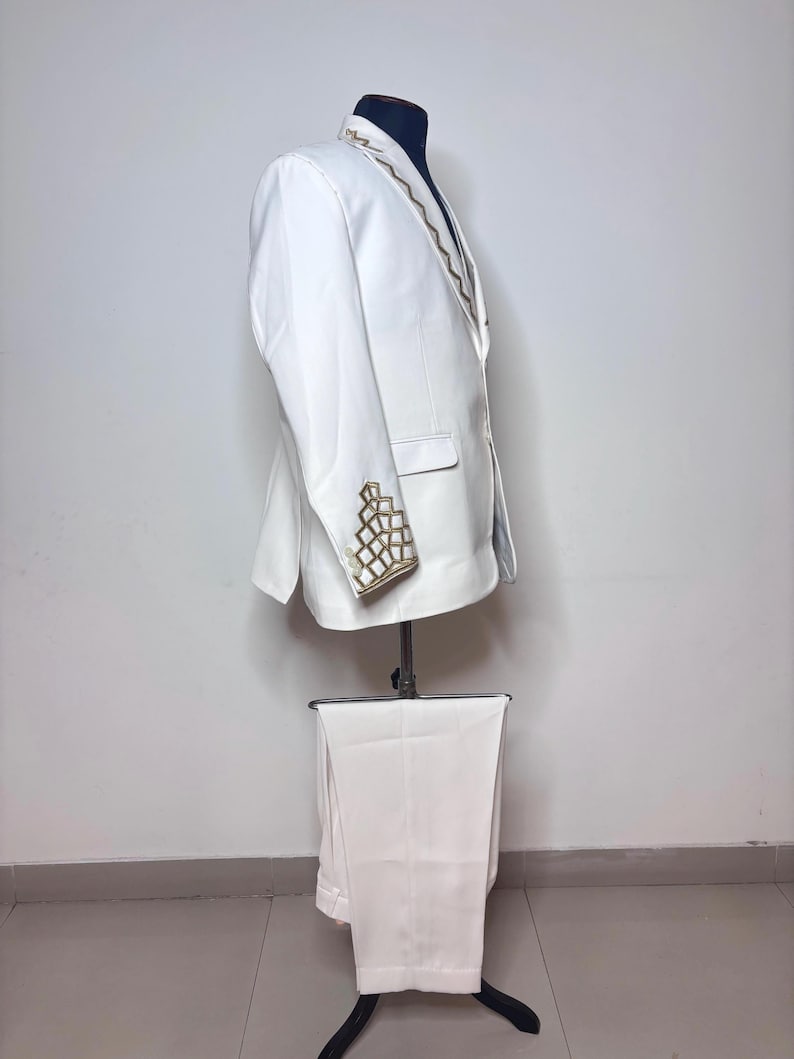 Puede incluir: Un traje blanco exhibido en un maniqu&iacute;. El traje incluye una chaqueta con bordados geom&eacute;tricos dorados en la solapa y los pu&ntilde;os, y pantalones a juego. El traje es entallado y parece estar hecho de una tela lisa.
