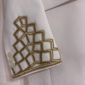 Puede incluir: Blazer blanco con bordado geom&eacute;trico dorado en la manga. El blazer tiene tres botones blancos en el pu&ntilde;o. El bordado dorado est&aacute; hecho de peque&ntilde;as cuentas y crea un dise&ntilde;o texturizado.