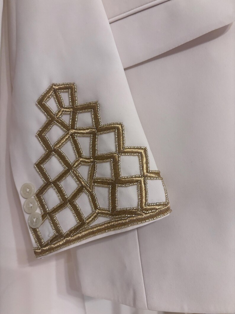 Puede incluir: Blazer blanco con bordado geom&eacute;trico dorado en la manga. El blazer tiene tres botones blancos en el pu&ntilde;o. El bordado dorado est&aacute; hecho de peque&ntilde;as cuentas y crea un dise&ntilde;o texturizado.