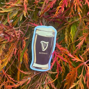 Puede incluir: Una pegatina que representa una pinta de Guinness con espuma blanca y un logotipo de arpa. La palabra "guinness" est&aacute; impresa debajo del arpa. La pegatina tiene un borde azul claro y est&aacute; sobre un fondo de hojas de oto&ntilde;o.