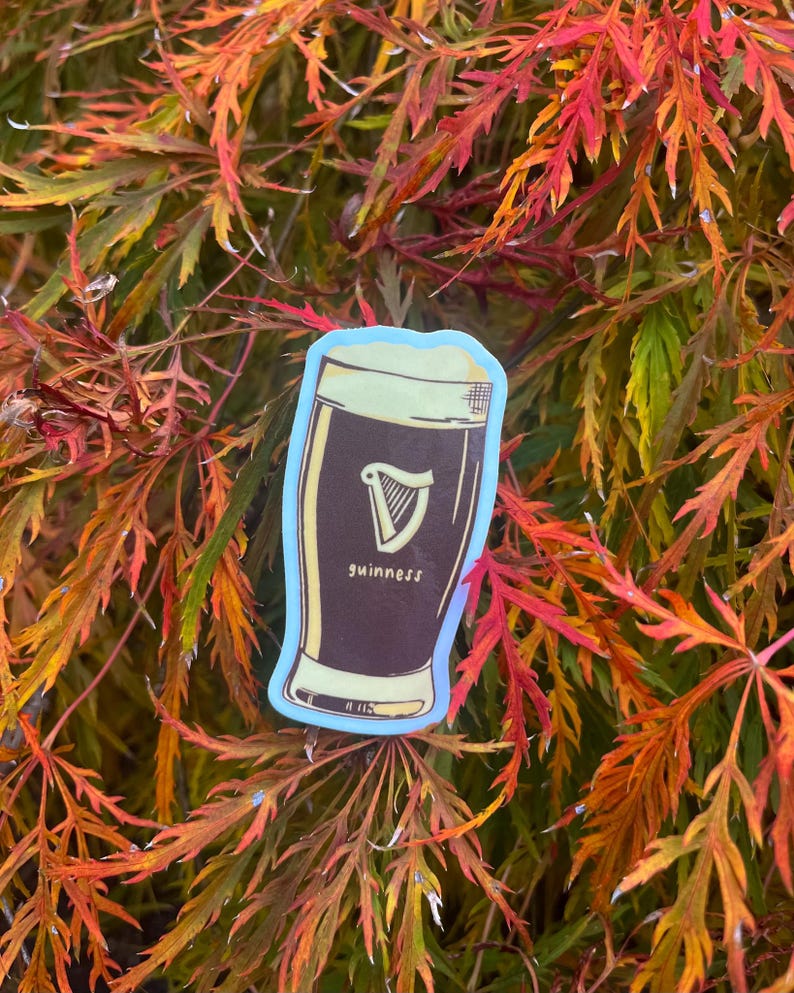 Puede incluir: Una pegatina que representa una pinta de Guinness con espuma blanca y un logotipo de arpa. La palabra "guinness" est&aacute; impresa debajo del arpa. La pegatina tiene un borde azul claro y est&aacute; sobre un fondo de hojas de oto&ntilde;o.
