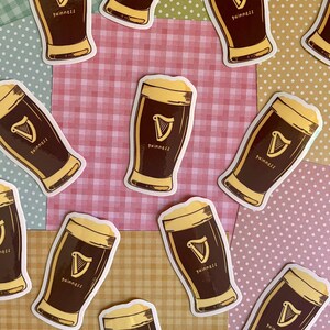 Puede incluir: Una colecci&oacute;n de pegatinas de cerveza Guinness. Cada pegatina presenta una pinta de cerveza oscura con espuma, el logotipo de Guinness y la palabra "Guinness". Las pegatinas est&aacute;n dispersas sobre un fondo de retazos de varios colores y patrones.