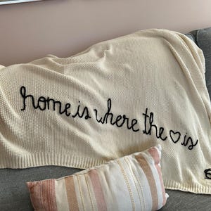 Könnte beinhalten: Eine cremefarbene Strickdecke mit dem in Schwarz gestickten Text "home is where the heart is", wobei ein Herzsymbol das Wort heart ersetzt. Die Decke hat ein strukturiertes Waffelstrickmuster und einen gerippten Rand an der Unterseite.