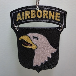 May include: Black keychain with a shield design featuring a white eagle head with a yellow beak and red tongue. The word "AIRBORNE" is in gold letters on a black banner above the shield. The keychain is attached to a silver chain.