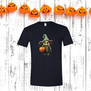 May include: A black t-shirt featuring a Halloween-themed graphic. The graphic depicts a cartoon creature wearing a witch hat and holding a jack-o'-lantern. Orange pumpkin lights hang above the shirt against a white wooden background.