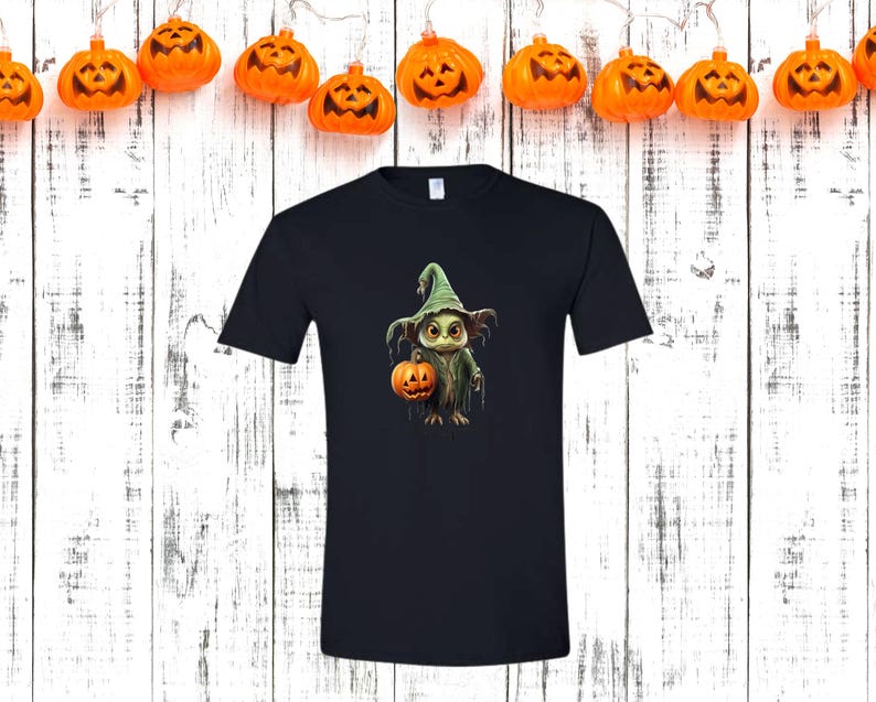 May include: A black t-shirt featuring a Halloween-themed graphic. The graphic depicts a cartoon creature wearing a witch hat and holding a jack-o'-lantern. Orange pumpkin lights hang above the shirt against a white wooden background.