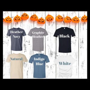 May include: A selection of t-shirts in various colors, including Heather Navy, Graphite Heather, Black, Natural, Indigo Blue, and White. Each shirt displays its color name in a contrasting font. The shirts are arranged against a white wooden background, with orange pumpkin decorations.