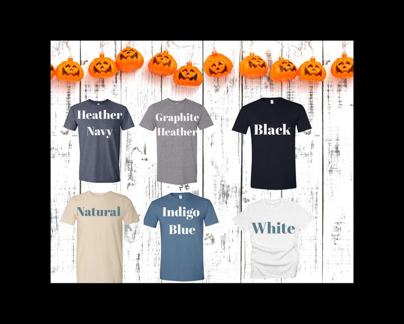 May include: A selection of t-shirts in various colors, including Heather Navy, Graphite Heather, Black, Natural, Indigo Blue, and White. Each shirt displays its color name in a contrasting font. The shirts are arranged against a white wooden background, with orange pumpkin decorations.