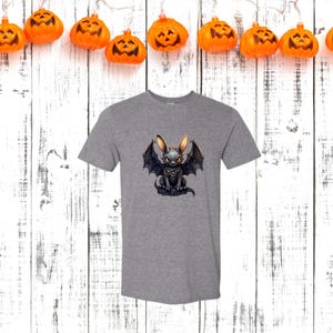 May include: A heather gray t-shirt featuring a cartoon bat-bunny hybrid with large ears and bat wings. The creature is black with yellow accents. The shirt is displayed against a white wooden background with orange pumpkin lights.
