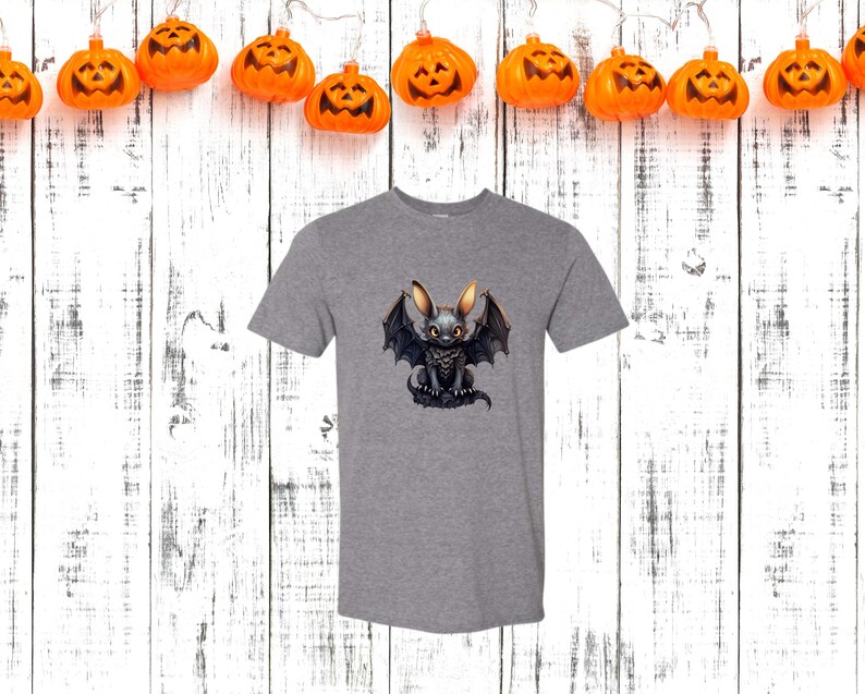 May include: A heather gray t-shirt featuring a cartoon bat-bunny hybrid with large ears and bat wings. The creature is black with yellow accents. The shirt is displayed against a white wooden background with orange pumpkin lights.