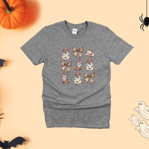 May include: A heather gray t-shirt featuring a repeating pattern of cowboy boots, bows, and hats. The design is in shades of brown and white. The shirt is surrounded by Halloween decorations, including pumpkins, spiders, bats, and ghosts.