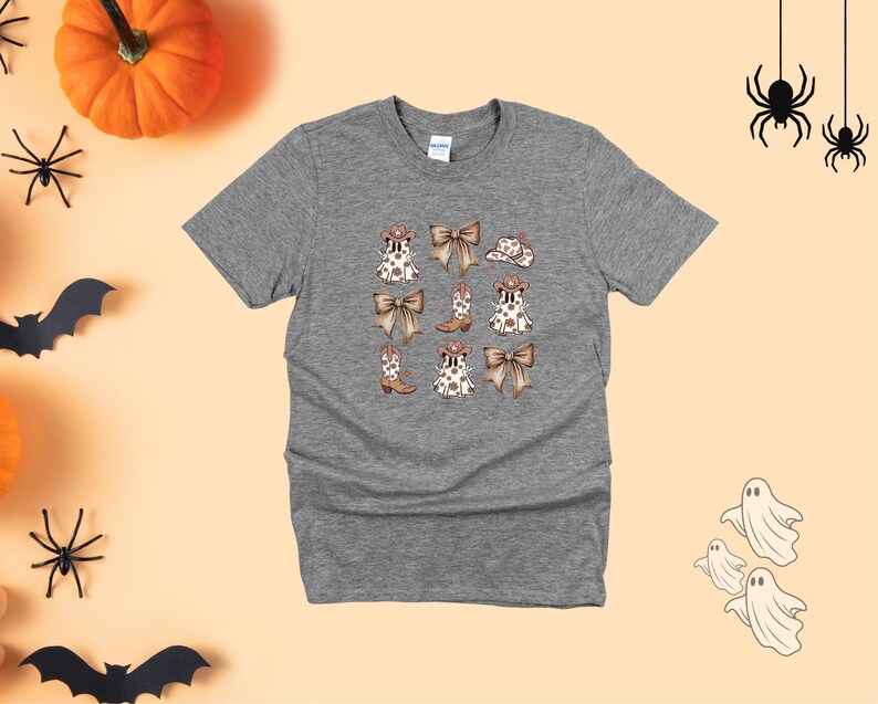 May include: A heather gray t-shirt featuring a repeating pattern of cowboy boots, bows, and hats. The design is in shades of brown and white. The shirt is surrounded by Halloween decorations, including pumpkins, spiders, bats, and ghosts.