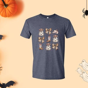 May include: A navy blue t-shirt featuring a repeating pattern of white and brown bows, cowboy boots, and hats. The design has a Halloween theme, with a pumpkin, spiders, bats, and ghosts in the background.