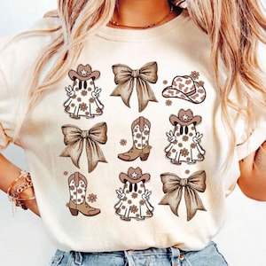 May include: Cream-colored t-shirt featuring a repeating pattern of cowboy ghost characters, bows, and cowboy boots in shades of brown and tan. The ghosts are wearing cowboy hats and have floral details.