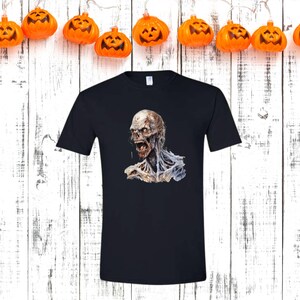 May include: Black t-shirt featuring a detailed illustration of a zombie with exposed flesh and an open mouth. The zombie graphic is in shades of gray, white, and red. The shirt is displayed against a white wooden background with orange pumpkin lights.