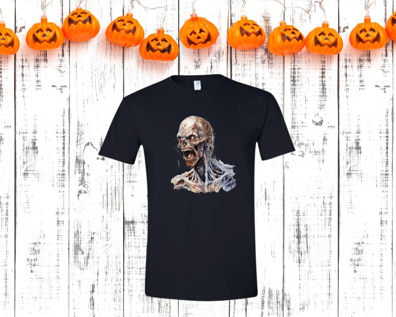 May include: Black t-shirt featuring a detailed illustration of a zombie with exposed flesh and an open mouth. The zombie graphic is in shades of gray, white, and red. The shirt is displayed against a white wooden background with orange pumpkin lights.