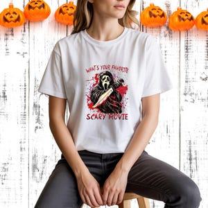 May include: White t-shirt featuring a graphic of a character from the movie Scream holding a knife, with the text "WHAT'S YOUR FAVORITE SCARY MOVIE" in red. The shirt is worn by a person sitting on a wooden stool. Orange pumpkin decorations are hanging in the background.
