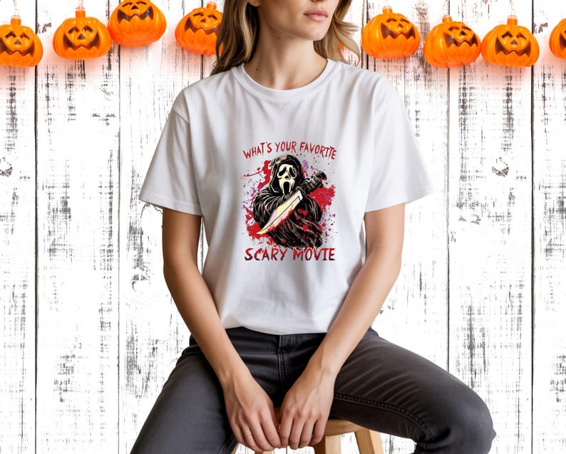 May include: White t-shirt featuring a graphic of a character from the movie Scream holding a knife, with the text "WHAT'S YOUR FAVORITE SCARY MOVIE" in red. The shirt is worn by a person sitting on a wooden stool. Orange pumpkin decorations are hanging in the background.