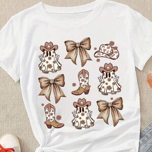 May include: White t-shirt featuring a repeating graphic of cartoon ghosts wearing cowboy hats and boots, with brown bows and floral accents. The design has a country western theme.