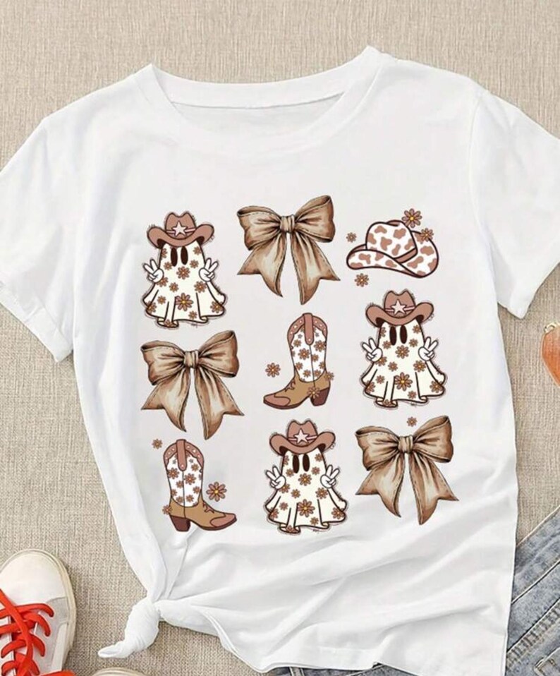 May include: White t-shirt featuring a repeating graphic of cartoon ghosts wearing cowboy hats and boots, with brown bows and floral accents. The design has a country western theme.