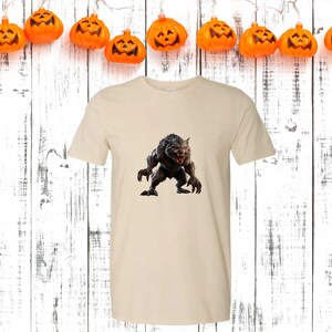 May include: A cream-colored t-shirt featuring a detailed graphic of a snarling werewolf. The werewolf is depicted in shades of gray and black, with sharp claws and teeth. The shirt is displayed against a backdrop of a white wooden surface and pumpkin lights, suggesting a Halloween theme.