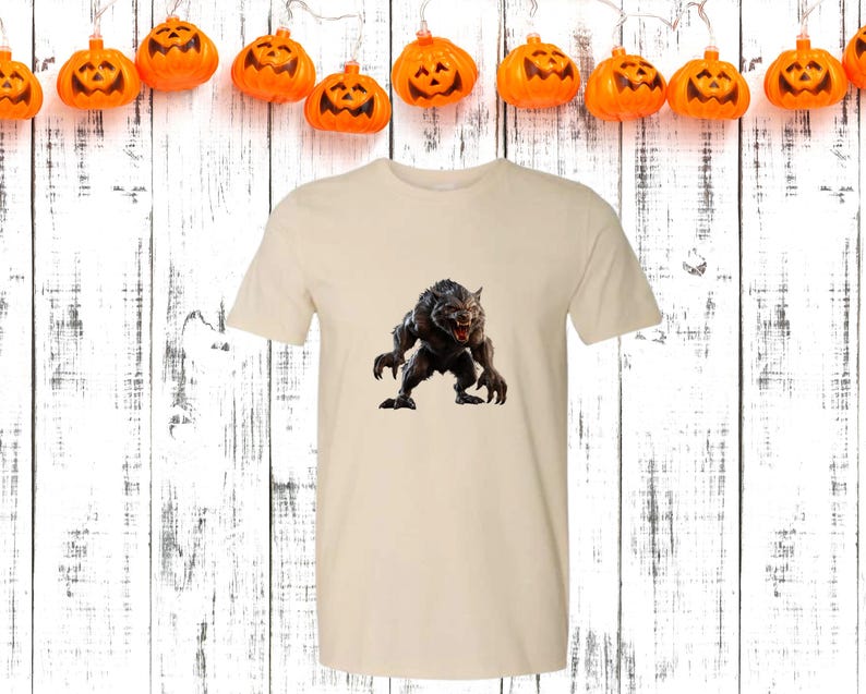 May include: A cream-colored t-shirt featuring a detailed graphic of a snarling werewolf. The werewolf is depicted in shades of gray and black, with sharp claws and teeth. The shirt is displayed against a backdrop of a white wooden surface and pumpkin lights, suggesting a Halloween theme.