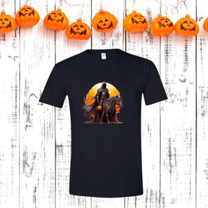 May include: Black t-shirt featuring a Halloween-themed graphic. The design depicts a cloaked figure on horseback holding a jack-o'-lantern, set against an orange sunset. The shirt is a classic crew neck style.
