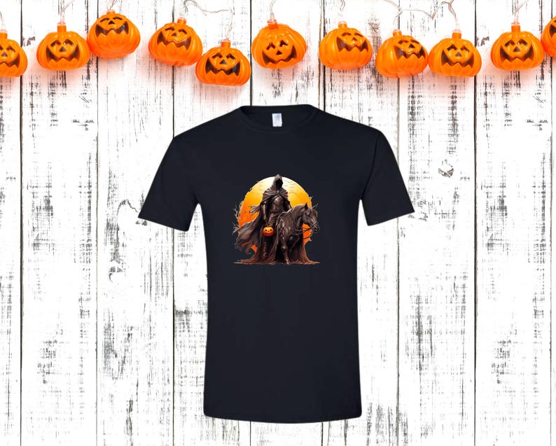 May include: Black t-shirt featuring a Halloween-themed graphic. The design depicts a cloaked figure on horseback holding a jack-o'-lantern, set against an orange sunset. The shirt is a classic crew neck style.
