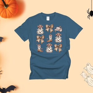May include: A teal t-shirt featuring a repeating pattern of cartoon ghosts wearing cowboy hats and boots, with brown bows. The design has a Halloween theme. The shirt is a short-sleeved crew neck.