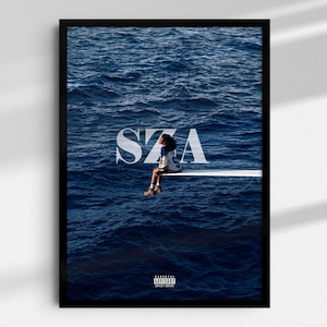May include: A framed wall art print featuring the SZA SOS album cover. The image displays a person sitting on a white diving board over deep blue ocean water. The large white text SZA is centered, and a small Parental Advisory label is at the bottom.