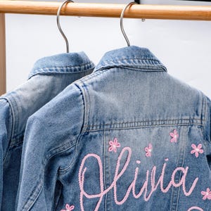 May include: Two light blue denim jackets hanging on a wooden rack. The jackets have pink embroidered floral accents and the name "Olivia" embroidered on the back. The jackets are made of denim.