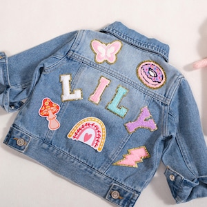 May include: A light blue denim jacket with colorful patches spelling "LILA". Patches include a butterfly, donut, mushroom, rainbow, lightning bolt, and the letter patches. A pink stuffed bunny is next to the jacket.