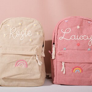 Könnte beinhalten: Zwei Rucksäcke aus Cord, einer beige und einer rosa, sind abgebildet. Der beige Rucksack hat den Namen "Rosie" in Weiß gestickt, mit Blumen- und Regenbogen-Details. Der rosa Rucksack zeigt den Namen "Laixy" und ähnliche Verzierungen.