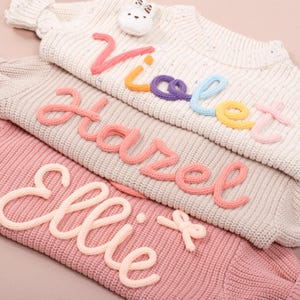 Könnte beinhalten: Drei Strickpullover in Creme, Grau und Rosa, jeder mit einem Namen, der mit farbigem Garn bestickt ist. Die Namen "Violet", "Hazel" und "Ellie" sind in einer verspielten Schreibschrift dargestellt. Ein kleines Häschen-Plüschtier befindet sich auf dem obersten Pullover.
