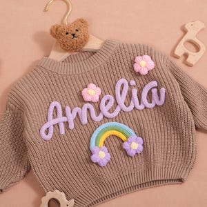 Può includere: Un maglione marrone lavorato a maglia con il nome "Amelia" in corsivo lavanda, decorato con fiori lavanda e un arcobaleno. Un appendiabiti a forma di orsetto e giocattoli di legno sono anche nell'immagine. Il maglione è per neonati o bambini piccoli.