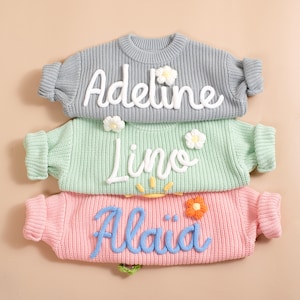 May include: Three pastel-coloured knit sweaters. The top sweater is grey with the name "Adeline" in white script and white flower accents. The middle sweater is mint green with "Lino" and a sun design. The bottom sweater is pink with "Alaia" in blue.