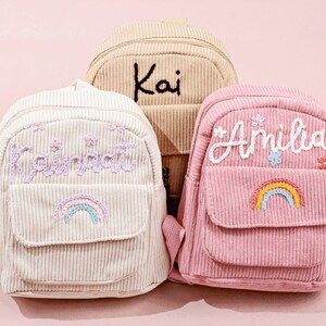 Könnte beinhalten: Drei Cord-Rucksäcke in Creme, Beige und Rosa. Jeder Rucksack hat eine Vordertasche und gestickte Details, darunter Namen und Regenbogen. Der cremefarbene Rucksack hat den Namen "Kainat" in Lavendel gestickt.