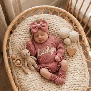May include: A dusty rose knitted long-sleeve romper featuring the name Evelyn embroidered in white script, accented with small bows. The outfit includes a matching headband with a large bow and knitted socks with bow details, paired with a crochet rabbit.