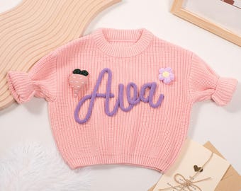 May include: A light pink knit sweater with the name "Ava" in purple script across the front. The sweater features a carrot and flower applique. The sleeves have a ruffled design. A card and wooden shapes are in the background.