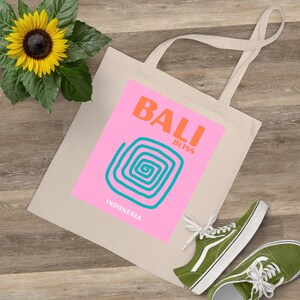 Op de afbeelding: Een beige draagtas met de woorden "BALI BLISS" in oranje en turquoise, met een turquoise spiraalontwerp op een roze achtergrond. Het woord "INDONESIA" staat eronder. Een zonnebloem en groene sneakers staan ook in de afbeelding.