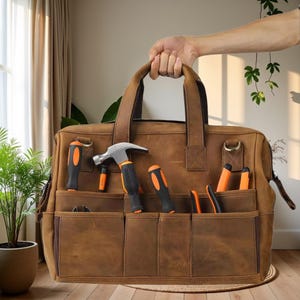 Könnte beinhalten: Eine braune Werkzeugtasche aus Leder mit mehreren Taschen, gefüllt mit verschiedenen Werkzeugen, darunter ein Hammer und Schraubendreher. Die Tasche hat einen Griff und wird hochgehalten. Die Werkzeuge haben schwarze und orangefarbene Griffe.