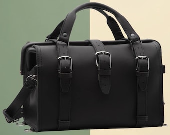 May include: Black satchel bag with a top handle and a detachable shoulder strap. The bag features three decorative straps with silver buckles on the front. The bag has a rectangular shape and is made of a smooth material.