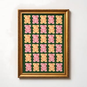 Puede incluir: Una impresión enmarcada que presenta un patrón de ositos de goma en tonos rosa, amarillo y naranja sobre un fondo verde oscuro. La impresión está encerrada en un marco dorado, agregando un toque de elegancia al diseño lúdico. Una obra de arte colorida.