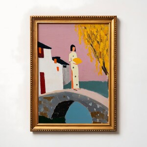 May include: Oil painting in a gold frame depicting a person in a white dress holding a yellow fan, standing on a stone bridge. A white building with a red door and orange accents is on the left. A yellow tree is on the right, with a pink sky.