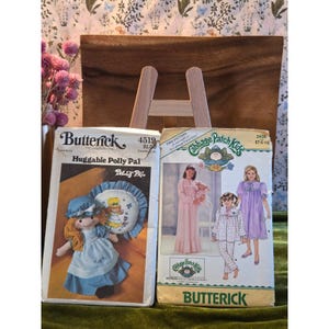 May include: Two vintage Butterick sewing patterns are displayed. One features a doll named "Huggable Polly Pal," and the other showcases "Cabbage Patch Kids" designs. The patterns are set against a wooden easel and a floral backdrop.