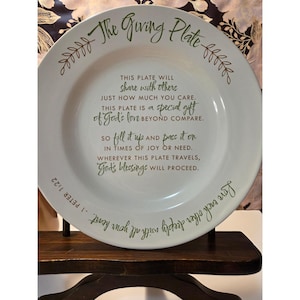 May include: White ceramic plate with the text "The Giving Plate" in green script. The plate includes a message about sharing and giving, with decorative leaf accents. The plate is displayed on a dark wooden stand.