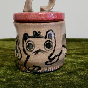 May include: A handmade ceramic jar with a lid. The jar is tan with a red lid and a curved handle. It features a whimsical cat design in black. The jar is likely used for storage or decoration.